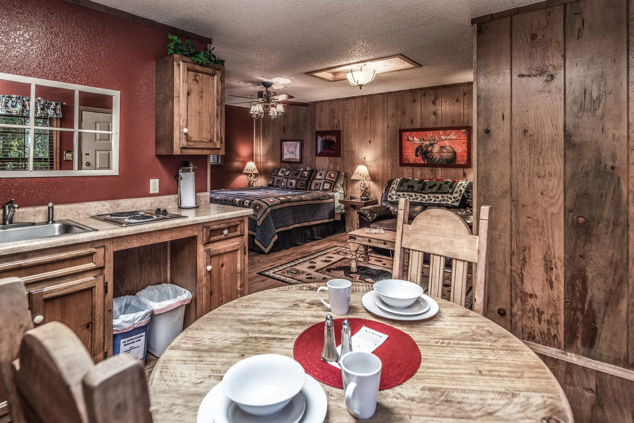 Cozy interior of a small rental cottage featuring a wooden dining table set with plates and cups, a compact kitchen area with wooden cabinetry, and a living space with a sofa, warm lighting, and wooden panel walls. The room has a rustic charm with decorative art pieces and a welcoming atmosphere.