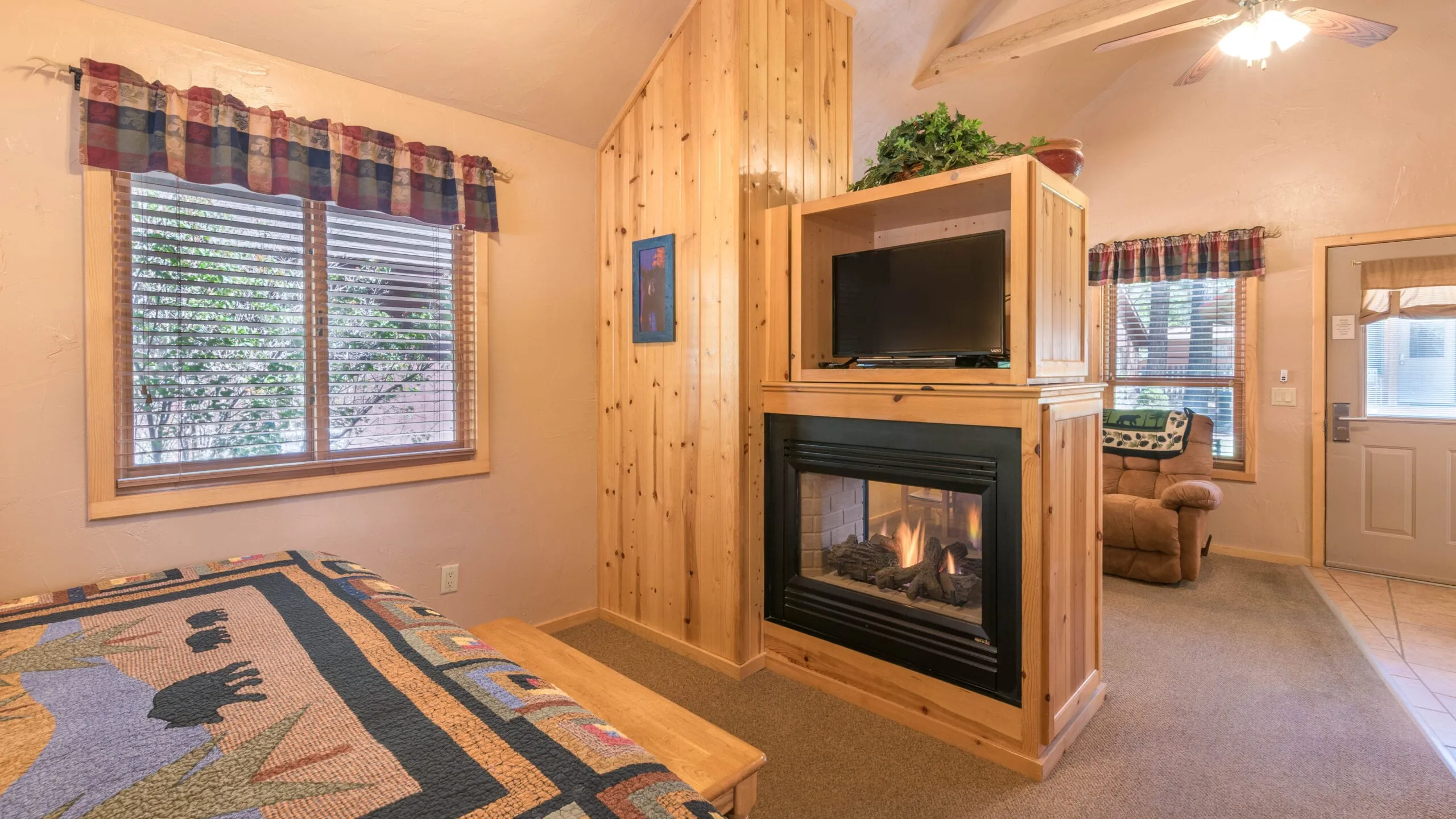 Cozy cabin interior featuring a wooden wall, a quilt-covered bed, a wall-mounted TV above a wooden fireplace, and a window with plaid curtains. Natural light fills the room, creating a warm atmosphere with a comfortable armchair in the corner.