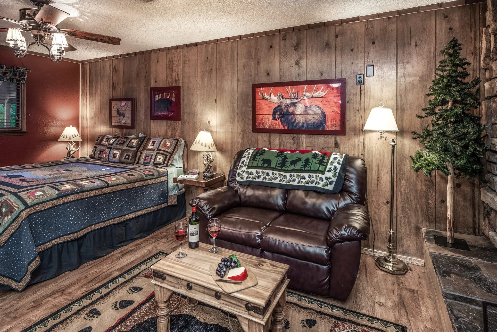 Cozy cabin bedroom interior featuring a king-size bed with a patterned quilt, a leather loveseat, wooden accent table, and rustic pine paneling. Two bedside lamps provide warm lighting, and a decorative moose painting is hung on the wall. A small evergreen tree adds a touch of nature to the space.