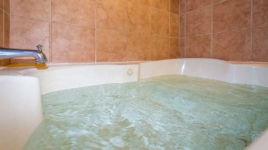 A close-up view of a filled corner bathtub with clear water, surrounded by beige tiled walls.
