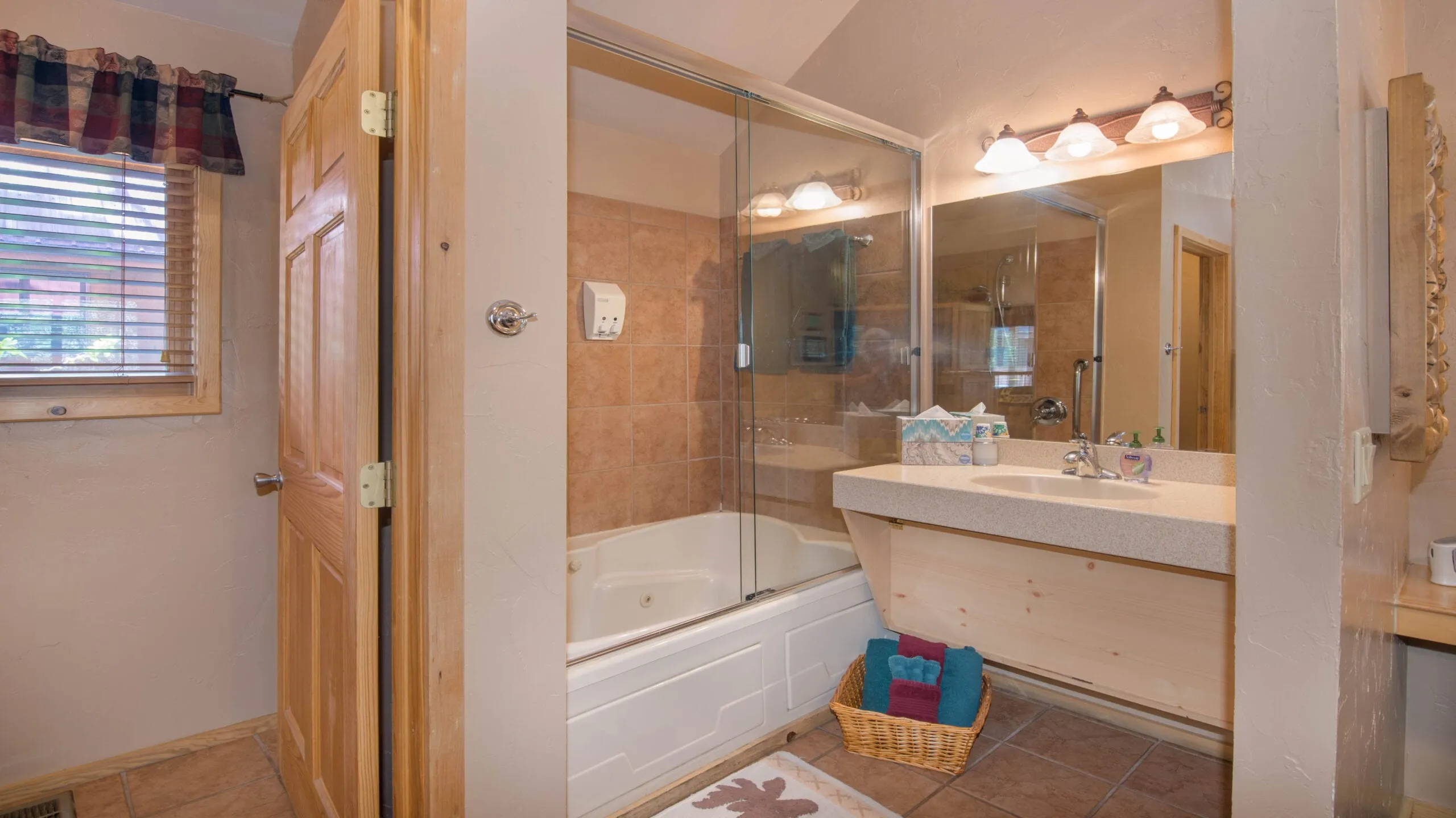 A modern bathroom featuring a glass shower enclosure, a white bathtub, and a double sink vanity with a mirror and light fixtures. The floor is tiled, and a wicker basket containing towels is visible. Natural light enters through a window with a plaid curtain. The walls are painted in neutral tones.