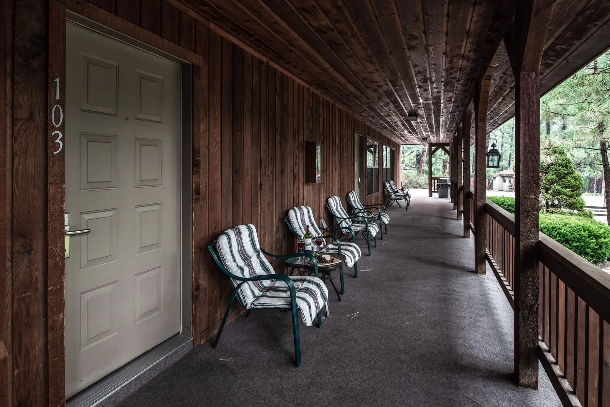A wooden corridor with a grey door labeled "103" on the left. Striped lounge chairs and small tables are lined up along the corridor. The surroundings feature dark wooden beams and lush greenery in the background, creating a serene atmosphere.