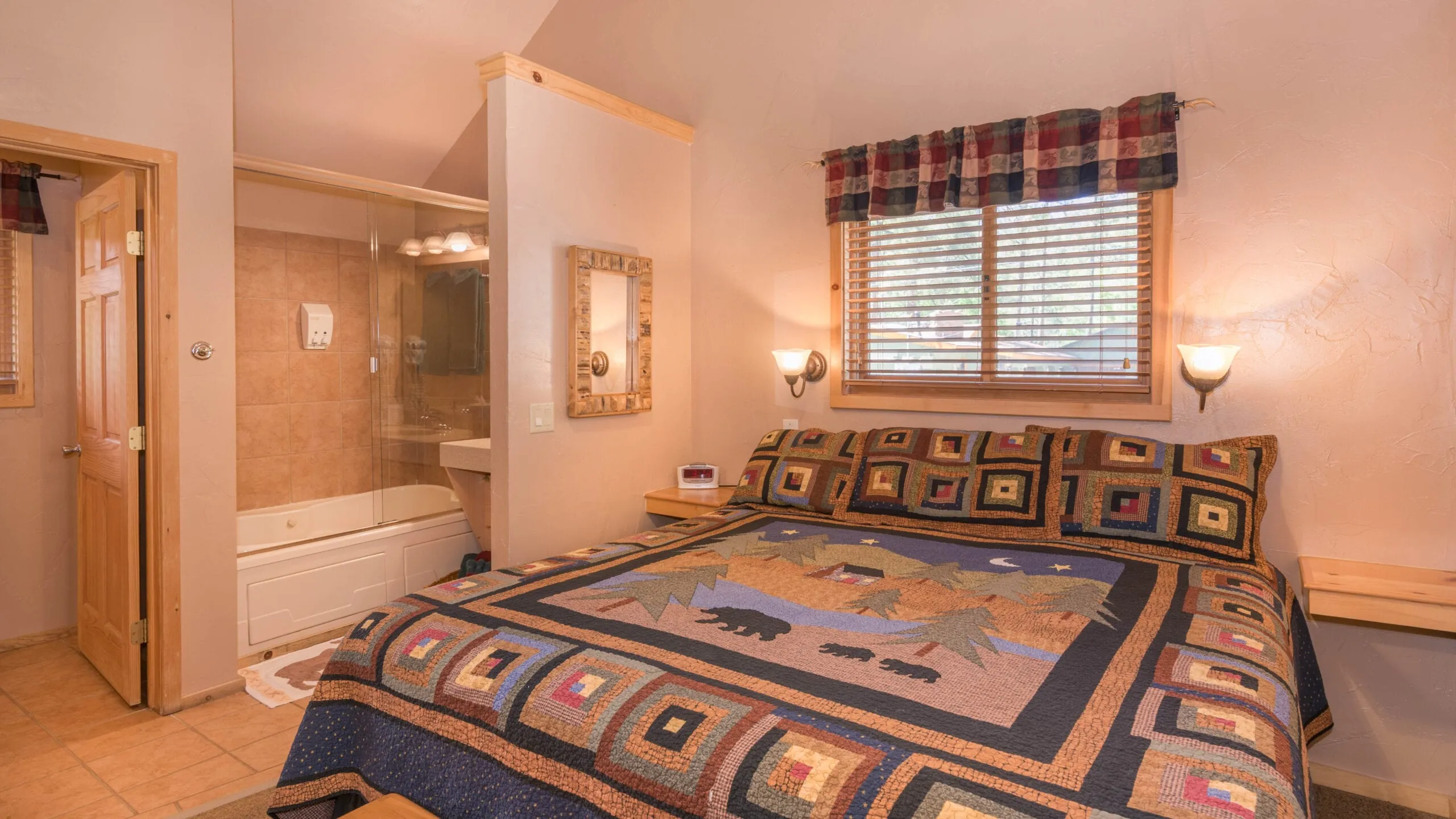 A cozy bedroom featuring a large bed adorned with a patterned quilt, two bedside lamps, and a window with blinds. A bathroom is partially visible in the background, with a mirror and light fixtures, indicating a warm and inviting atmosphere.