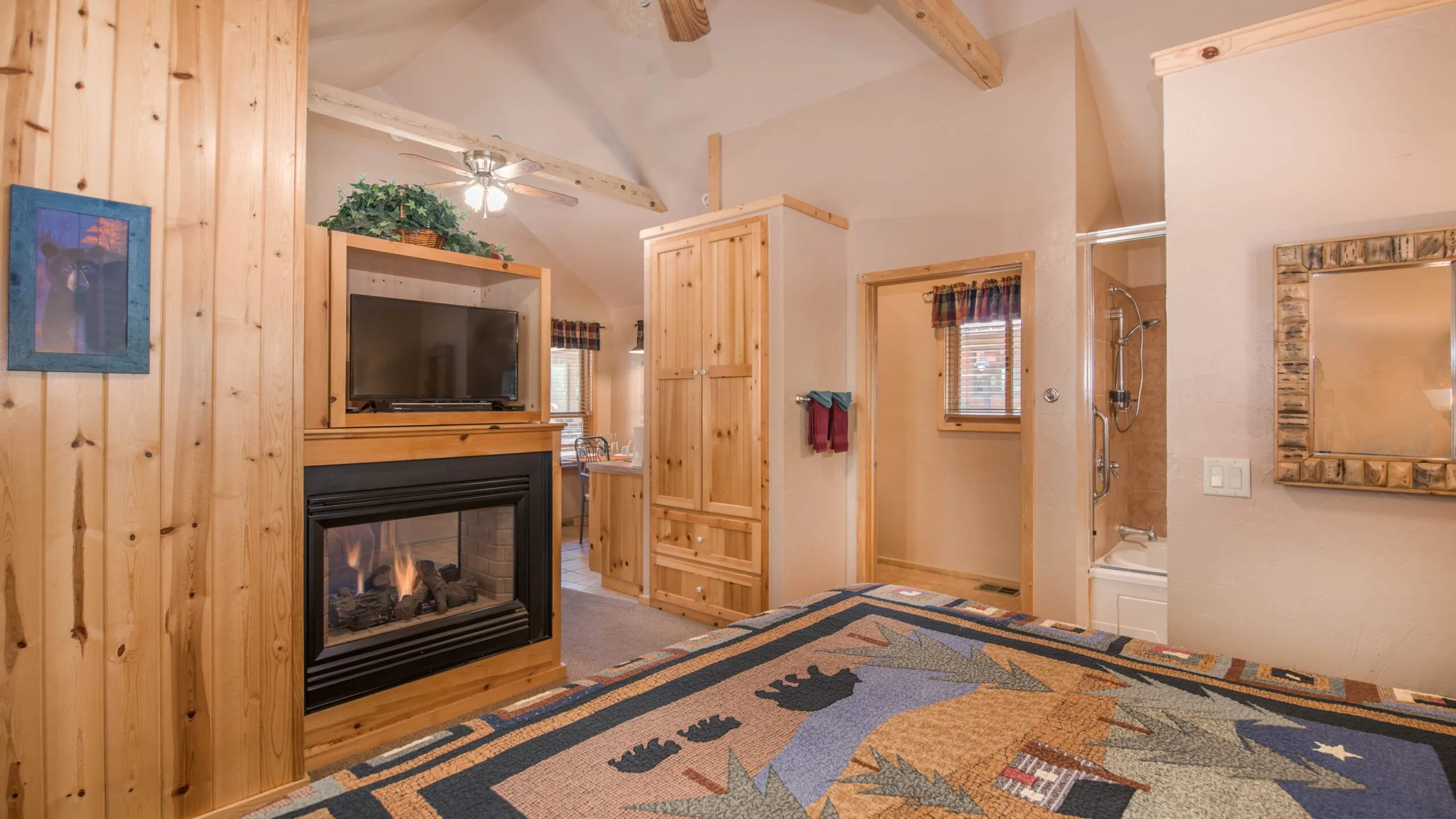Interior of a cozy cabin room featuring wooden accents, a fireplace, a television, and a large bed with a colorful quilt. The room includes a bathroom visible through an open doorway and has a warm, inviting ambiance.