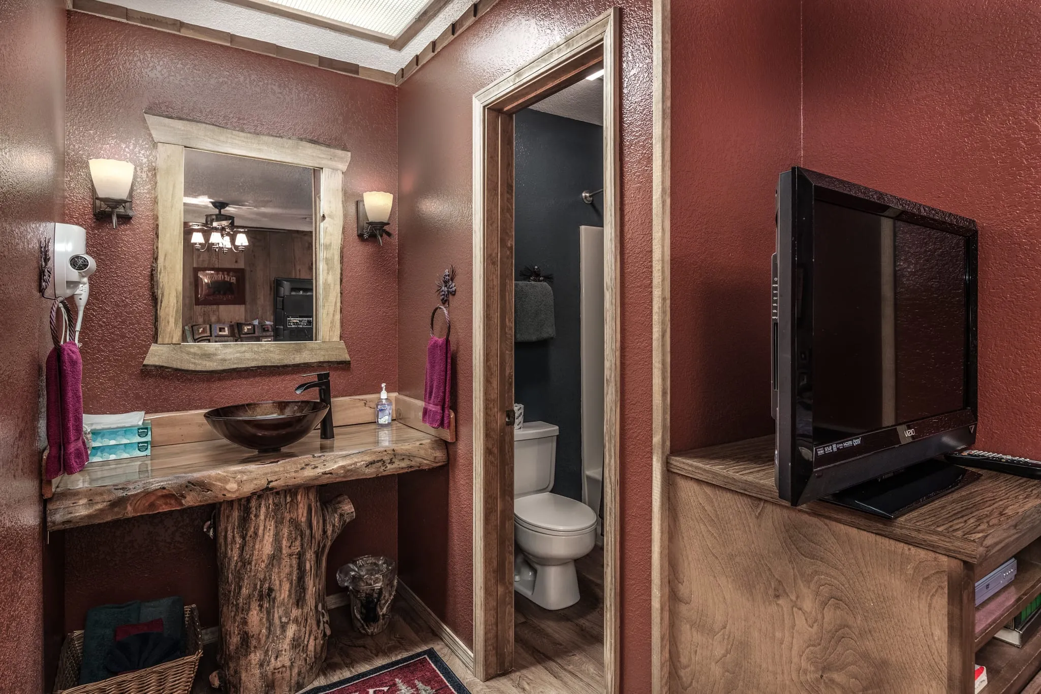 A cozy bathroom featuring a rustic wooden vanity with a round sink, a large mirror framed in wood, and adorned with decorative items. The walls are painted a warm reddish-brown, illuminated by wall sconces. To the left, a white toilet is visible through an open door, and a television is placed on a wooden shelf to the right. A patterned rug adds texture to the floor, enhancing the inviting atmosphere.