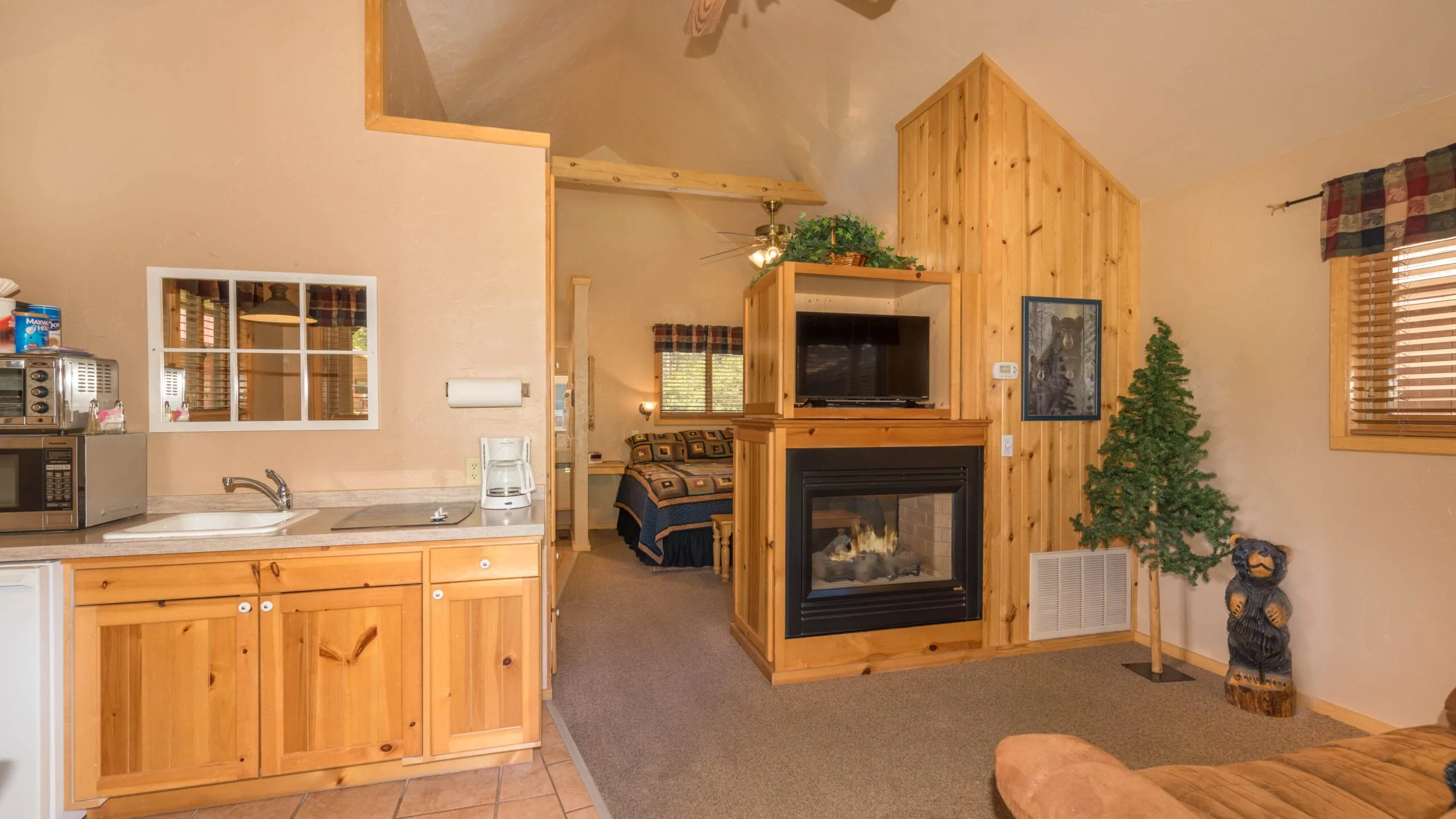 Interior view of a cozy cabin-style living room featuring wooden cabinetry, a fireplace, and a comfortable seating area. The space has high ceilings, large windows, and decorative elements like a small Christmas tree, creating a warm and inviting atmosphere.