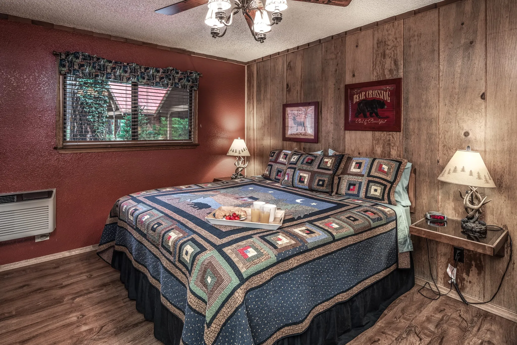 Cozy bedroom featuring a large quilted bedspread in earthy tones, adorned with patterned throw pillows. Natural wood paneling on the walls adds warmth, complemented by a rustic-style fan hanging from the ceiling. Two bedside lamps illuminate small wooden side tables with decorative accents. A window with a patterned curtain lets in natural light, revealing a glimpse of greenery outside. The flooring is a warm-toned laminate, enhancing the inviting atmosphere of the room.