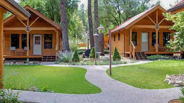 A scenic view of two wooden cabins surrounded by lush greenery and tall trees. A pathway made of gravel winds between the cabins, leading through a well-maintained lawn with shrubs and decorative plants. Sunlight filters through the trees, creating a tranquil and inviting atmosphere.