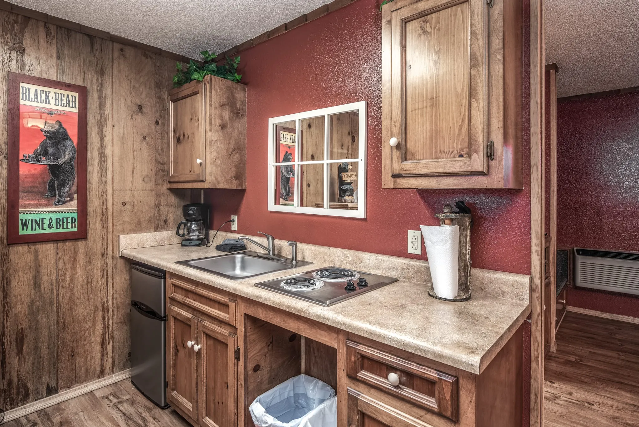Cozy kitchenette with wooden cabinets and countertops, featuring a small stovetop, sink, coffee maker, and refrigerator. A framed window looks out to a wall with rustic decor, including a bear-themed poster. The warm red walls add a homely atmosphere to the space.
