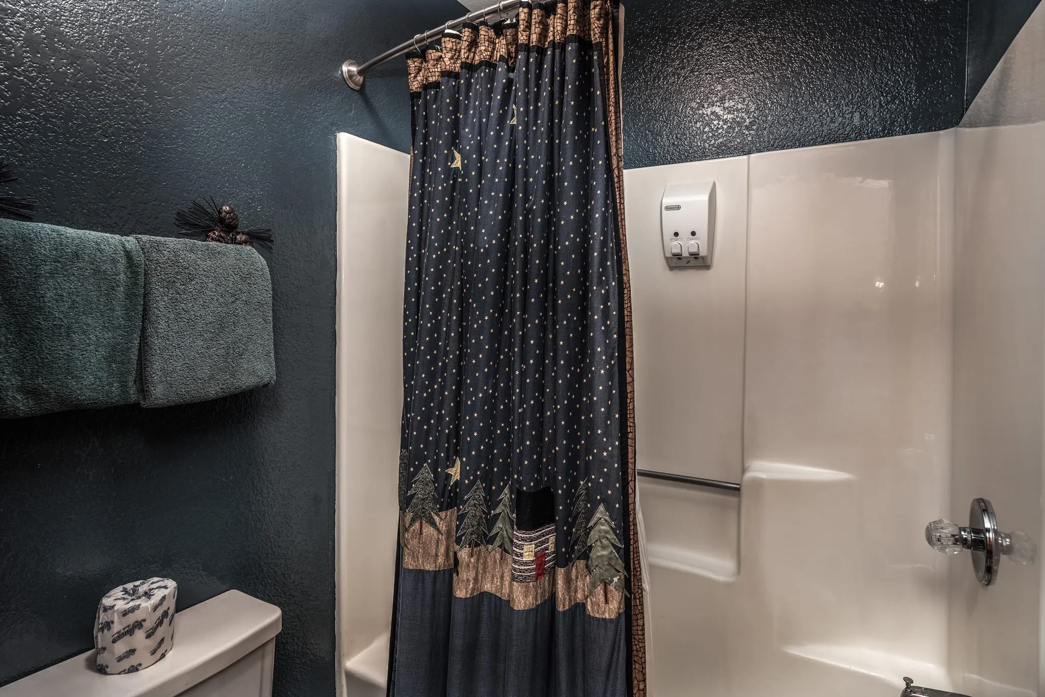 A modern bathroom featuring a shower with a dark patterned curtain, a light-colored bathtub, and a wall-mounted showerhead. There are neatly arranged towels on a rail and a roll of toilet paper on a nearby shelf. The walls are painted in a dark color, contributing to a cozy atmosphere.