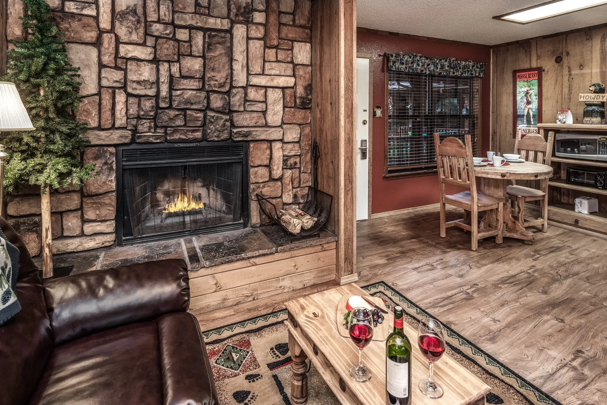Cozy living room featuring a stone fireplace, brown leather sofa, wooden coffee table with wine bottles and glasses, and a dining area with wooden chairs. The decor includes a small Christmas tree and a warm ambiance, creating an inviting atmosphere.