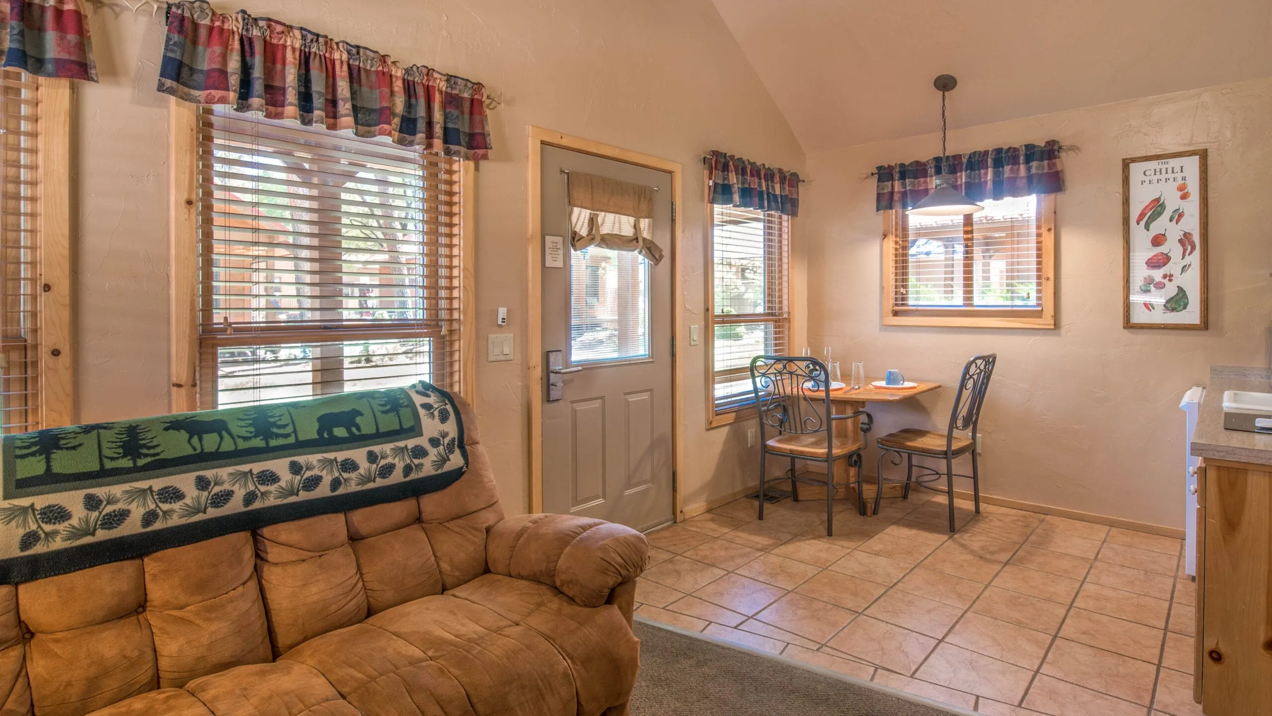 Interior view of a cozy living space featuring a tan leather couch, a small dining table with metal chairs, and large windows adorned with plaid curtains. The walls are painted in a warm tone, and a door is visible leading outside. A framed picture hangs on the wall, contributing to the homey atmosphere.