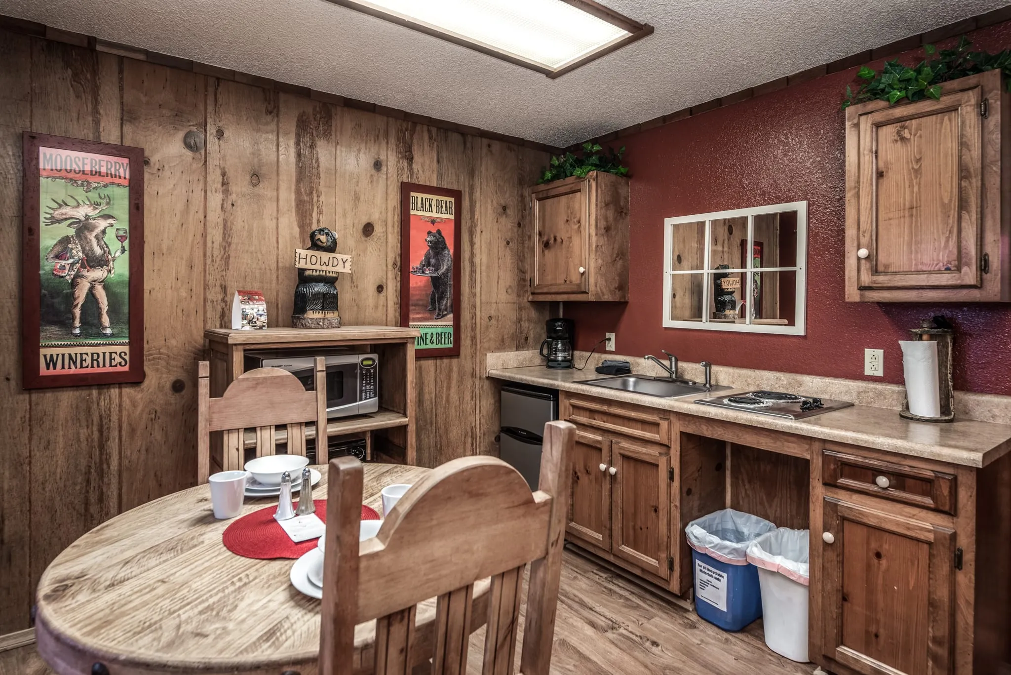 A cozy kitchen featuring wooden paneling and cabinets, a round table set for dining with plates and cutlery, a refrigerator, microwave, and stove, accented by decorative posters on the walls and greenery above the cabinets. The space is well-lit with a ceiling light and has a casual, rustic ambiance.