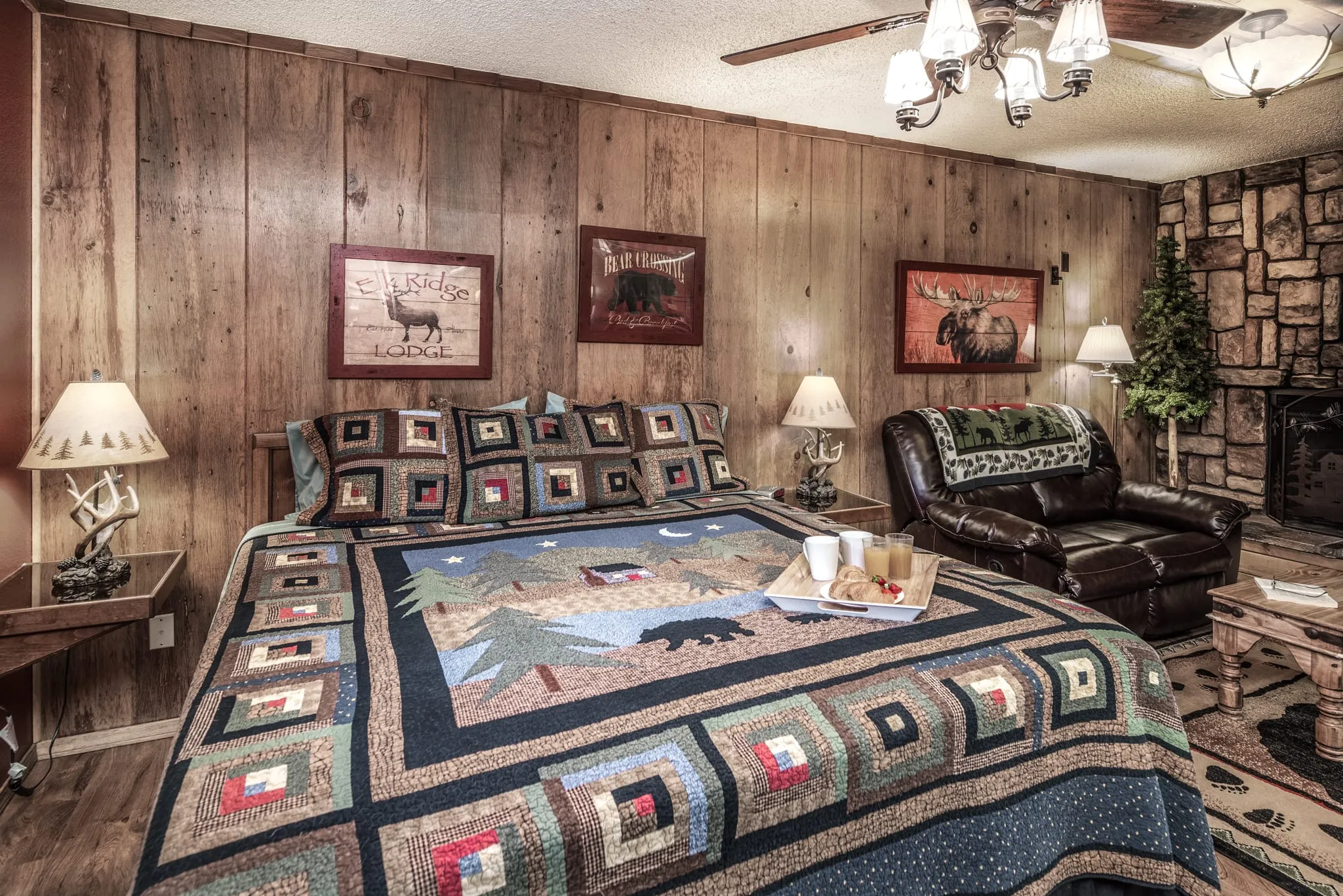 A cozy bedroom featuring a king-sized bed adorned with a colorful quilt displaying a nature design, complemented by wooden paneling on the walls. The room includes two bedside lamps, framed wildlife artwork, a plush leather sofa, and a stone fireplace. A small tray with snacks and beverages sits on the bed, enhancing the inviting atmosphere.