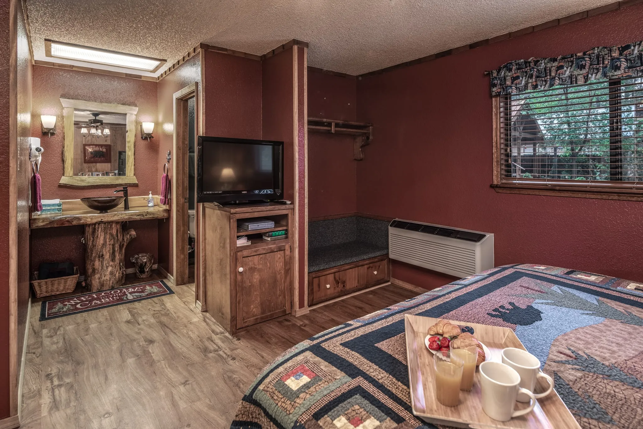 Cozy hotel room featuring a wooden bed with a patterned quilt, a small table with two cups and a snack, a TV on a wooden dresser, and natural light coming through a window with a plaid curtain. The walls are painted in a warm tone, and there is a small bench and a mirror on the wall.